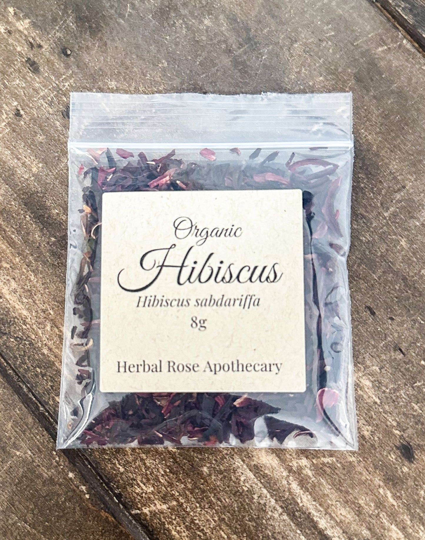 A 8g clear plastic resealable pouch of Organic Hibiscus tea by Herbal Rose Apothecary, featuring a cream-colored label, placed on a rustic wooden surface.