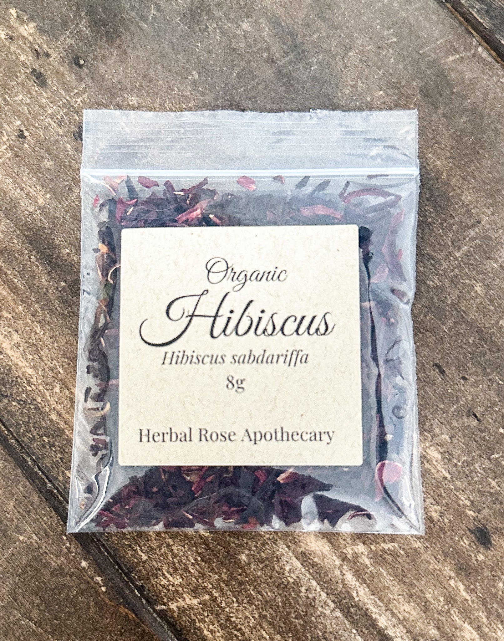 A 8g clear plastic resealable pouch of Organic Hibiscus tea by Herbal Rose Apothecary, featuring a cream-colored label, placed on a rustic wooden surface.