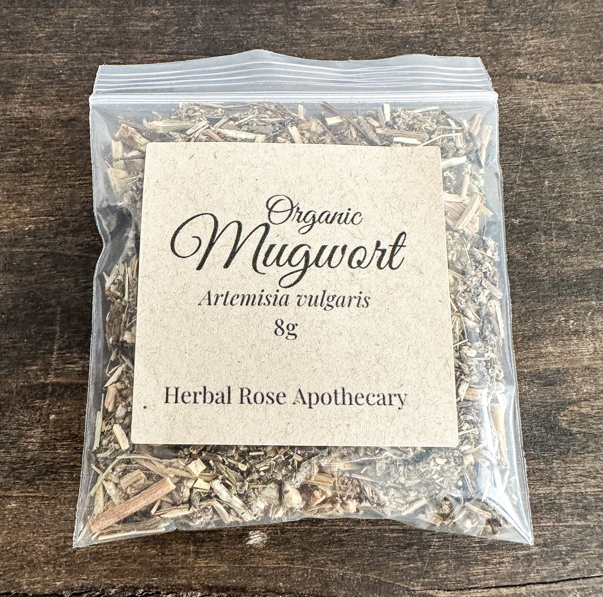 A clear plastic zip-top bag filled with dried mugwort herb, featuring a square kraft paper label that reads 'Organic Mugwort Artemisia vulgaris 8g' and 'Herbal Rose Apothecary,' placed on a dark wood surface.