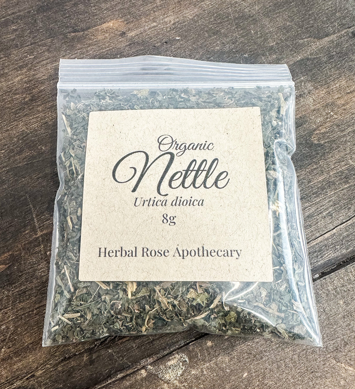 A clear plastic zip-top bag containing dried nettle leaves, with a beige paper label reading 'Organic Nettle Urtica dioica 8g Herbal Rose Apothecary,' placed on a dark wooden surface.