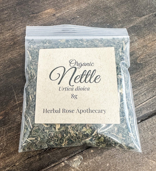 A clear plastic zip-top bag containing dried nettle leaves, with a beige paper label reading 'Organic Nettle Urtica dioica 8g Herbal Rose Apothecary,' placed on a dark wooden surface.