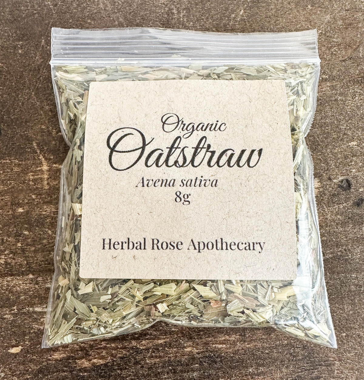 A clear plastic bag filled with dried oatstraw herbal tea, featuring a square kraft paper label that reads 'Organic Oatstraw Avena sativa 8g Herbal Rose Apothecary', set against a dark rustic wooden background.