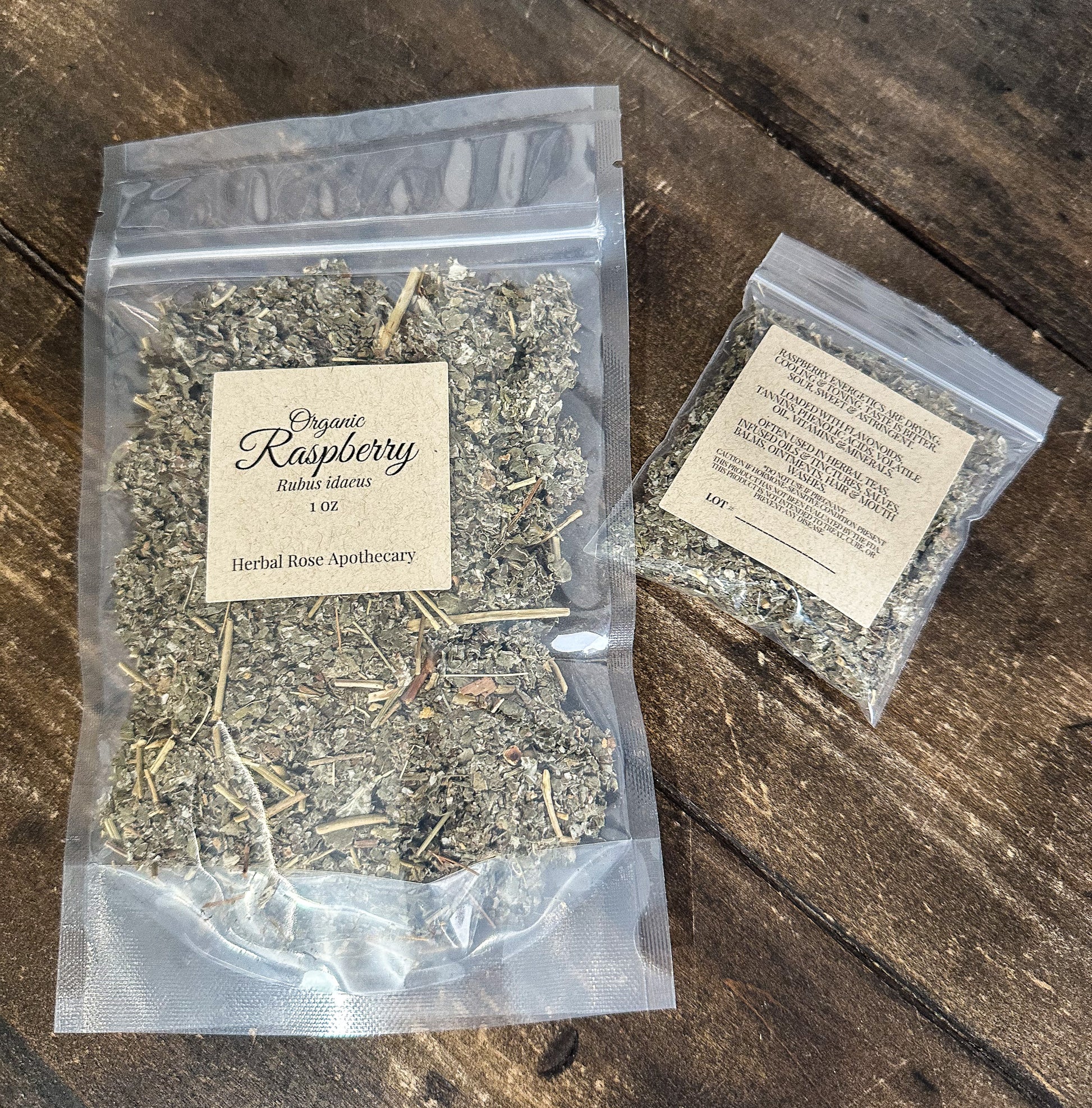 Two clear plastic bags of dried organic raspberry  on a dark rustic wooden surface. The larger bag on the left has a cream paper label reading 'Organic Raspberry' and the smaller bag on the right has a similar label with product text.