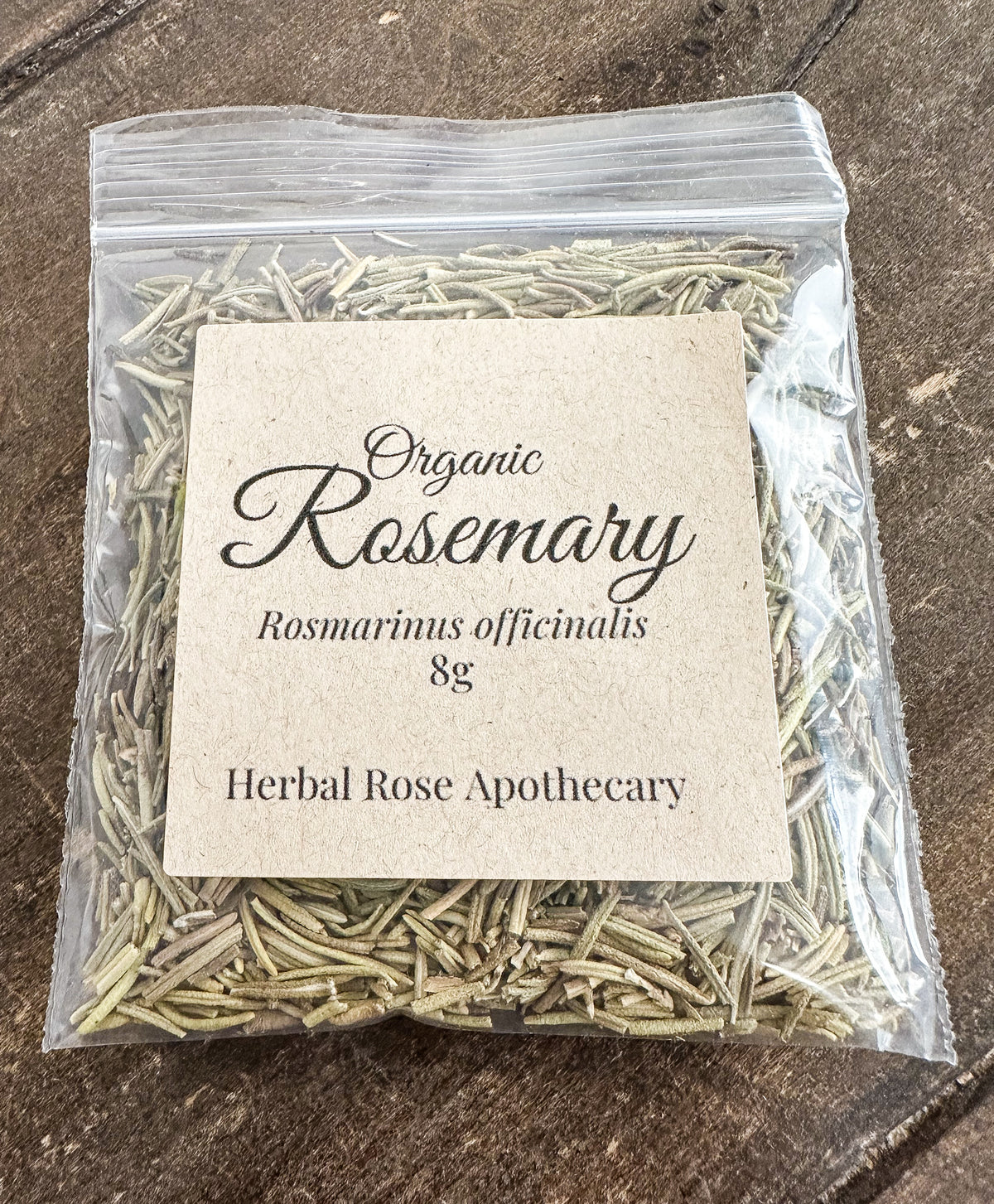 A clear plastic zip-top bag filled with dried rosemary stems and a large kraft paper label reading 'Organic Rosemary Rosmarinus officinalis 8g Herbal Rose Apothecary,' photographed on a dark wood surface.