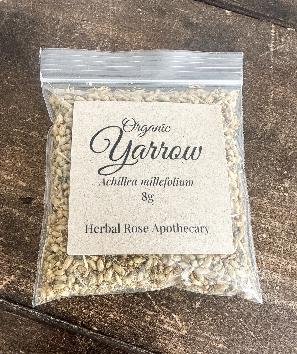A clear plastic zip-top bag filled with dried herbal seeds or powder, featuring a square kraft paper label that reads 'Organic Yarrow' and 'Achillea millefolium 8g', placed on a dark wooden surface.