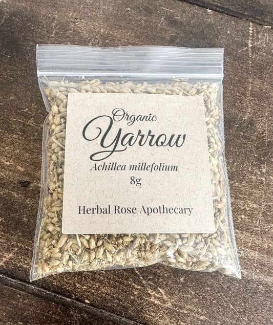 A clear plastic zip-top bag filled with dried herbal seeds or powder, featuring a square kraft paper label that reads 'Organic Yarrow' and 'Achillea millefolium 8g', placed on a dark wooden surface.