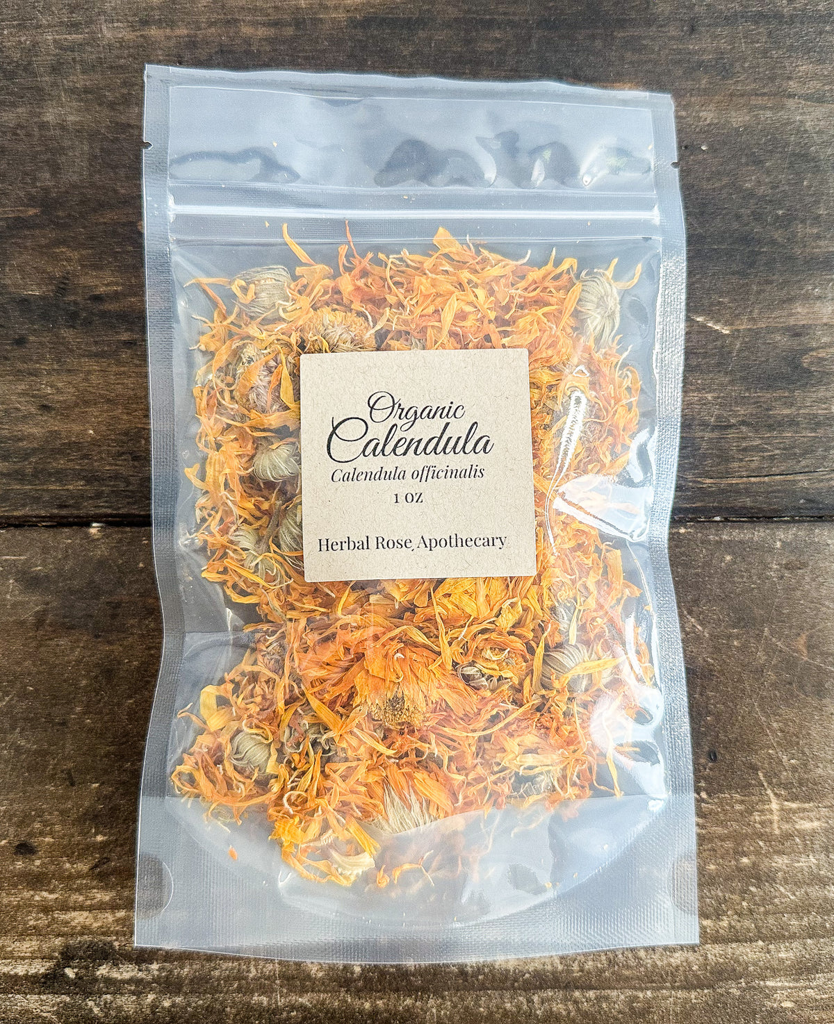 A clear resealable stand-up pouch containing dried orange calendula flowers and small bees, with a beige label reading 'Organic Calendula' and 'Herbal Rose Apothecary,' placed on a dark wooden surface.