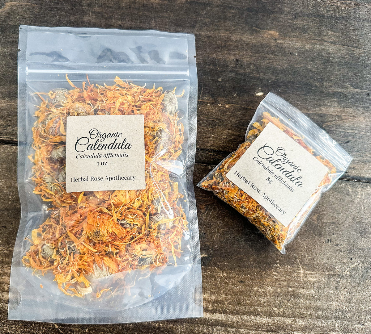 Two clear resealable pouches of Organic Calendula flowers from Herbal Rose Apothecary, displayed on a dark rustic wooden surface. One larger pouch contains 100 flowers and the smaller one contains 50, both showing the bright orange dried petals.