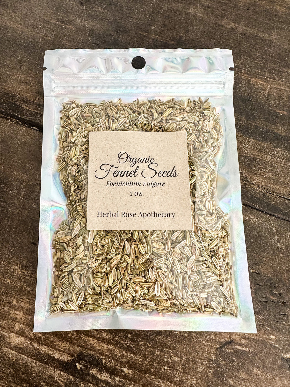 A 1 oz holographic stand-up pouch of Organic Fenel Seeds, displayed on a dark wood background. The pouch contains light brown seeds and features a beige paper label with black text.