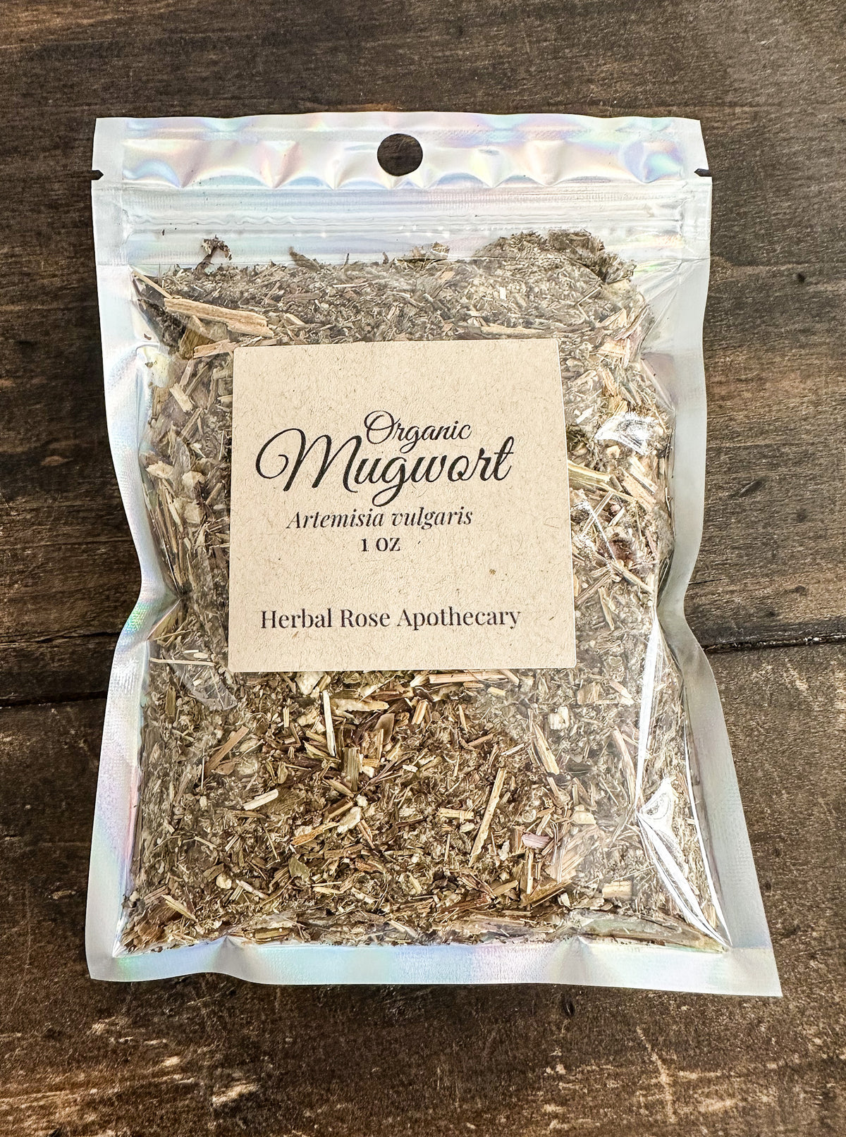 A 1 oz resealable stand-up pouch of Organic Mugwort Herbal Rose Apothecary, containing dried mugwort, set on a dark rustic wooden background.