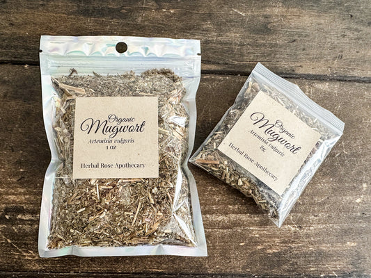 Two clear resealable pouches of Organic Mugwort herbal tea by Herbal Rose Apothecary, displayed on a dark rustic wooden background.
