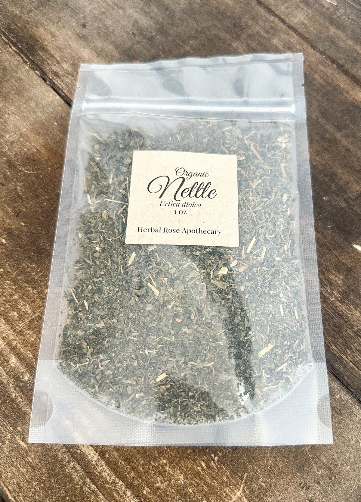 A clear resealable pouch containing dried herbal nettle tea, with a cream label reading 'Organic Nettle' and 'Herbal Rose Apothecary,' placed on a rustic wooden surface.