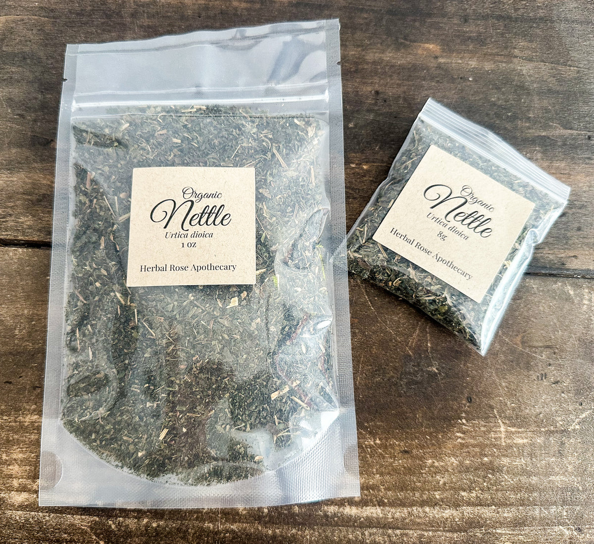 Two clear resealable pouches of Organic Nettle Herbal Rose Apothecary tea placed on a rustic dark wood surface. One large pouch and one small pouch, both with cream labels, showing the dried green herbal blend inside.