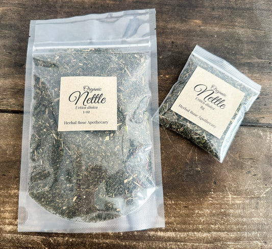 Two clear resealable pouches of Organic Nettle Herbal Rose Apothecary tea placed on a rustic dark wood surface. One large pouch and one small pouch, both with cream labels, showing the dried green herbal blend inside.