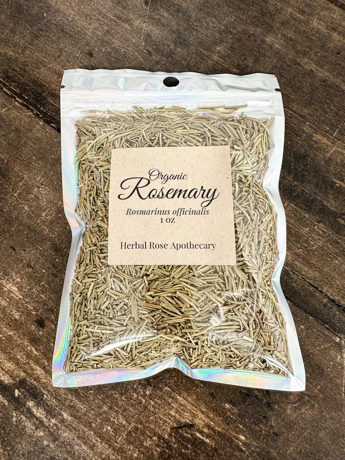 A 1 oz bag of Organic Rosemary from Herbal Rose Apothecary, featuring dried rosemary inside a clear holographic stand-up pouch with a beige paper label, set on a dark wooden surface.