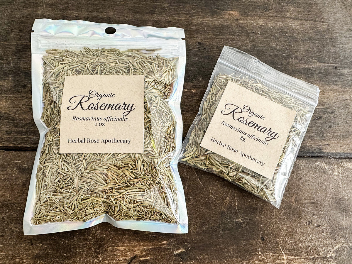 Two resealable bags of organic rosemary herb from Herbal Rose Apothecary, displayed on a dark rustic wooden surface. One larger bag is labeled 1 oz and one smaller bag is labeled 8 oz.