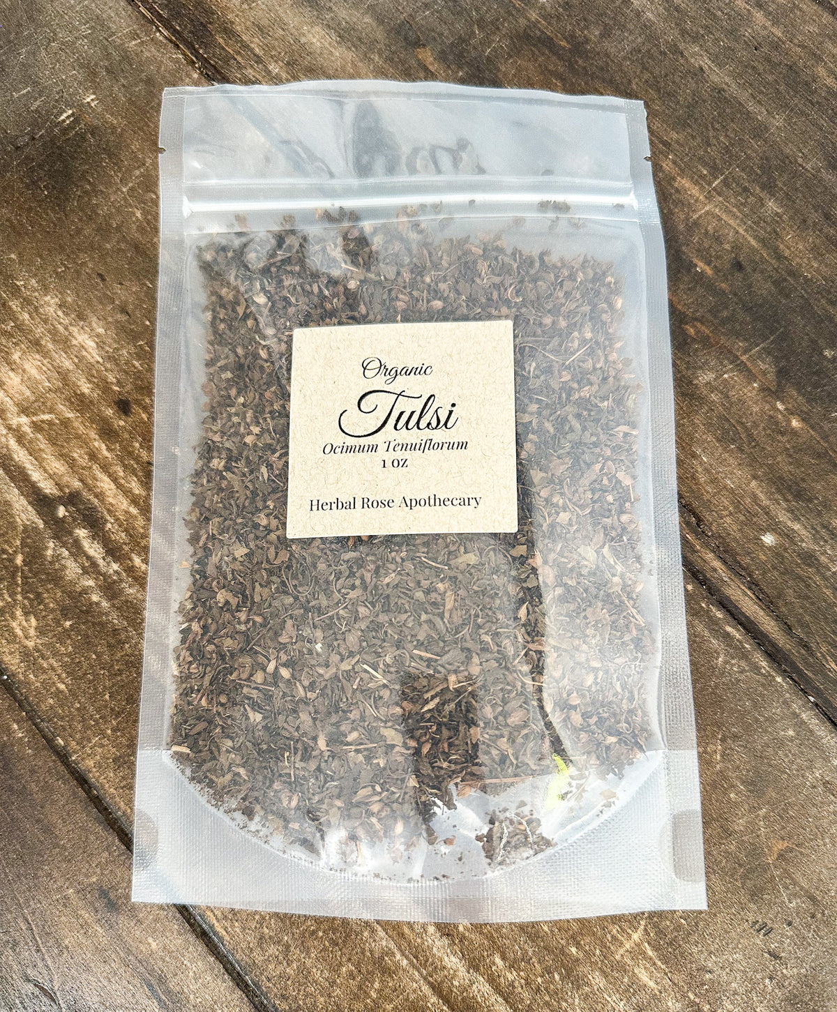 A clear stand-up pouch containing loose herbal tea, with a cream label reading 'Organic Tulsi' and 'Octenium Tranquillum 1 oz', placed on a rustic wooden surface.