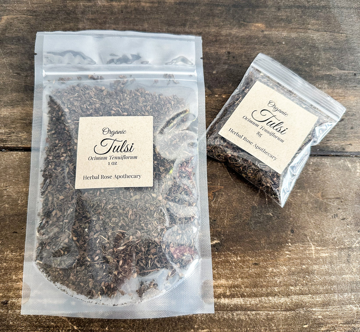Two clear resealable bags of Herbal Rose Apothecary Organic Tulsi tea placed on a rustic wooden surface. The larger bag in front shows the product name and brand on a cream label, and a smaller matching bag sits to the right.