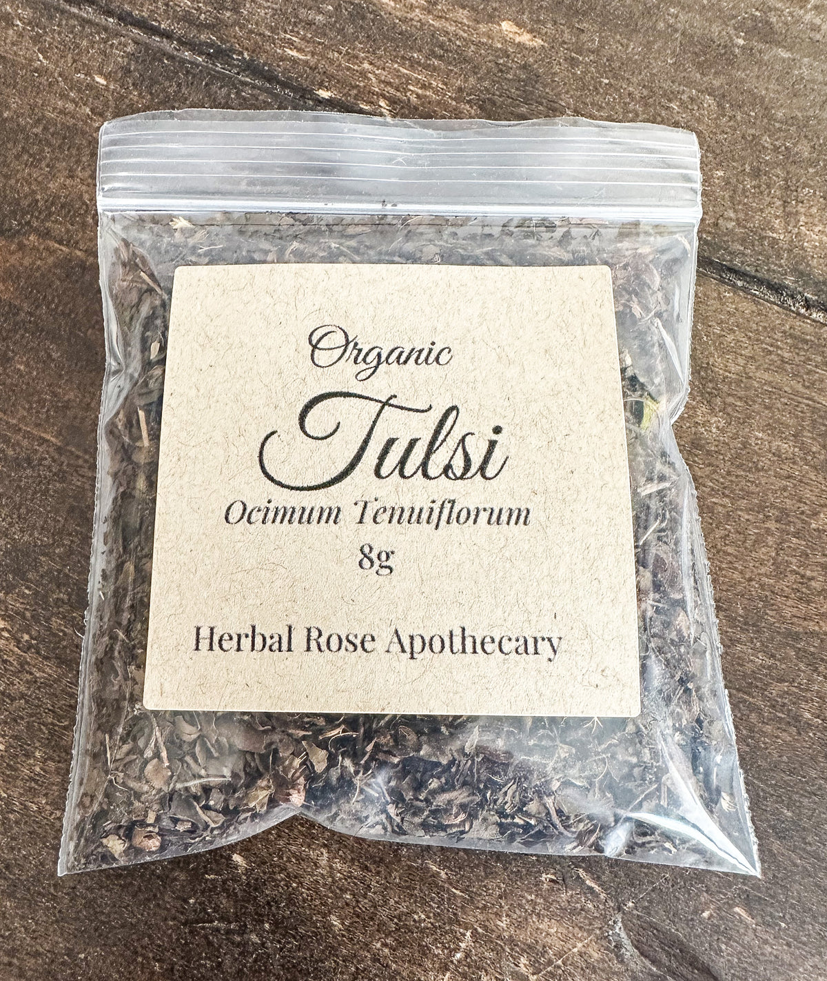 A clear plastic resealable pouch containing loose herbal tea, with a centered kraft paper label reading 'Organic Tulsi Ocimum Tenuiflorum 8g Herbal Rose Apothecary,' placed on a rustic wooden surface.