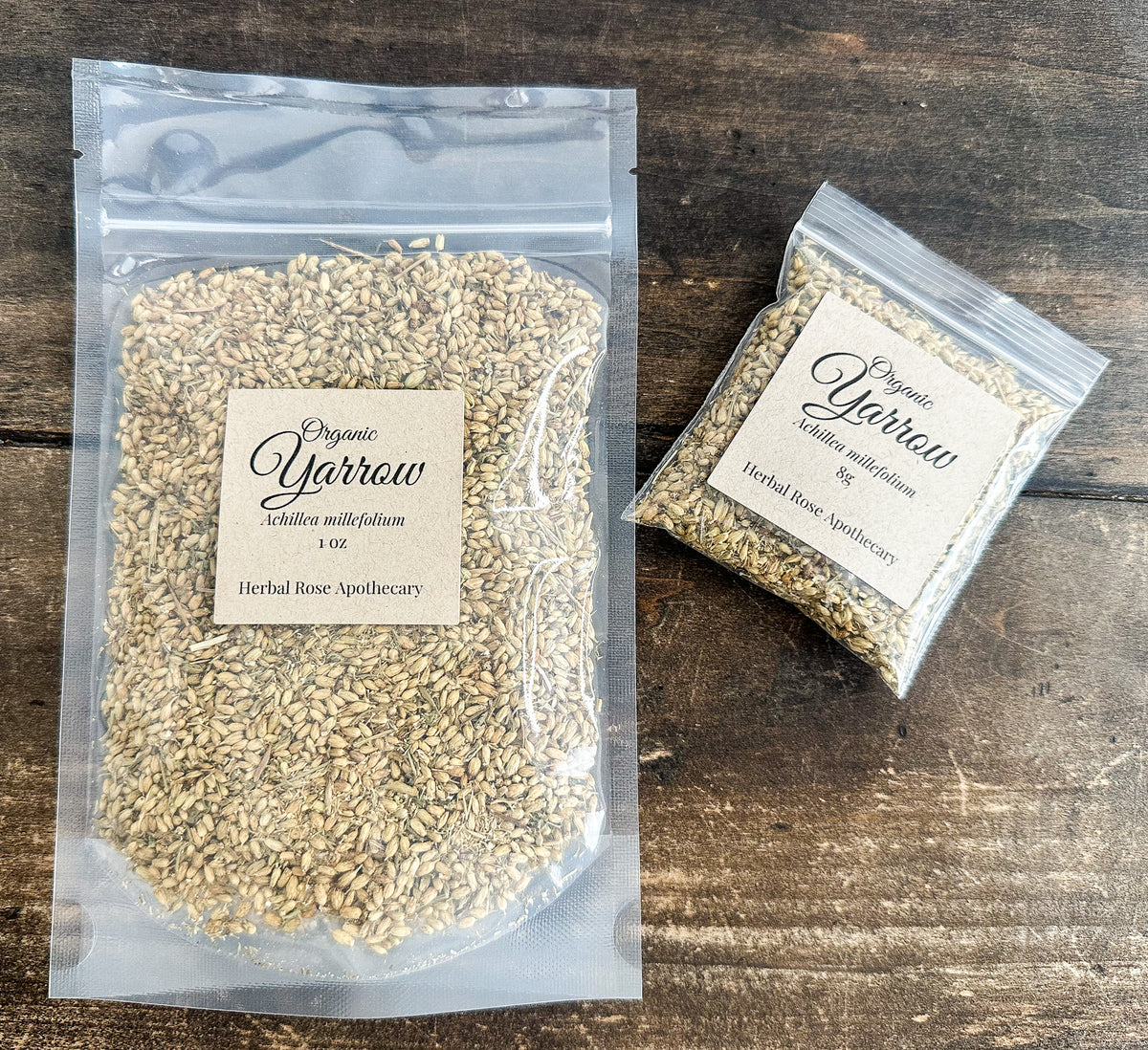 Two clear plastic bags of Organic Yarrow herbal rose apothecary seeds, one large 1 oz bag and one smaller 5 g bag, displayed on a dark rustic wooden surface.