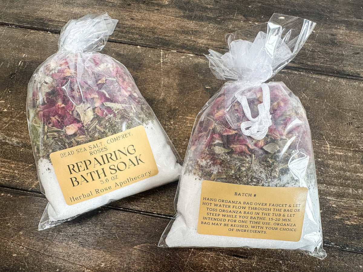 two repairing bath soaks packaged in a clear bag and labeled lying on a wooden surface from herbal rose apothecary