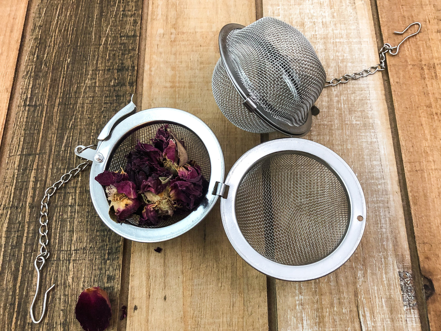 dried roses spilling out of an open tea ball infuser with an additional tea ball infuser next to it closed on a wooden surface