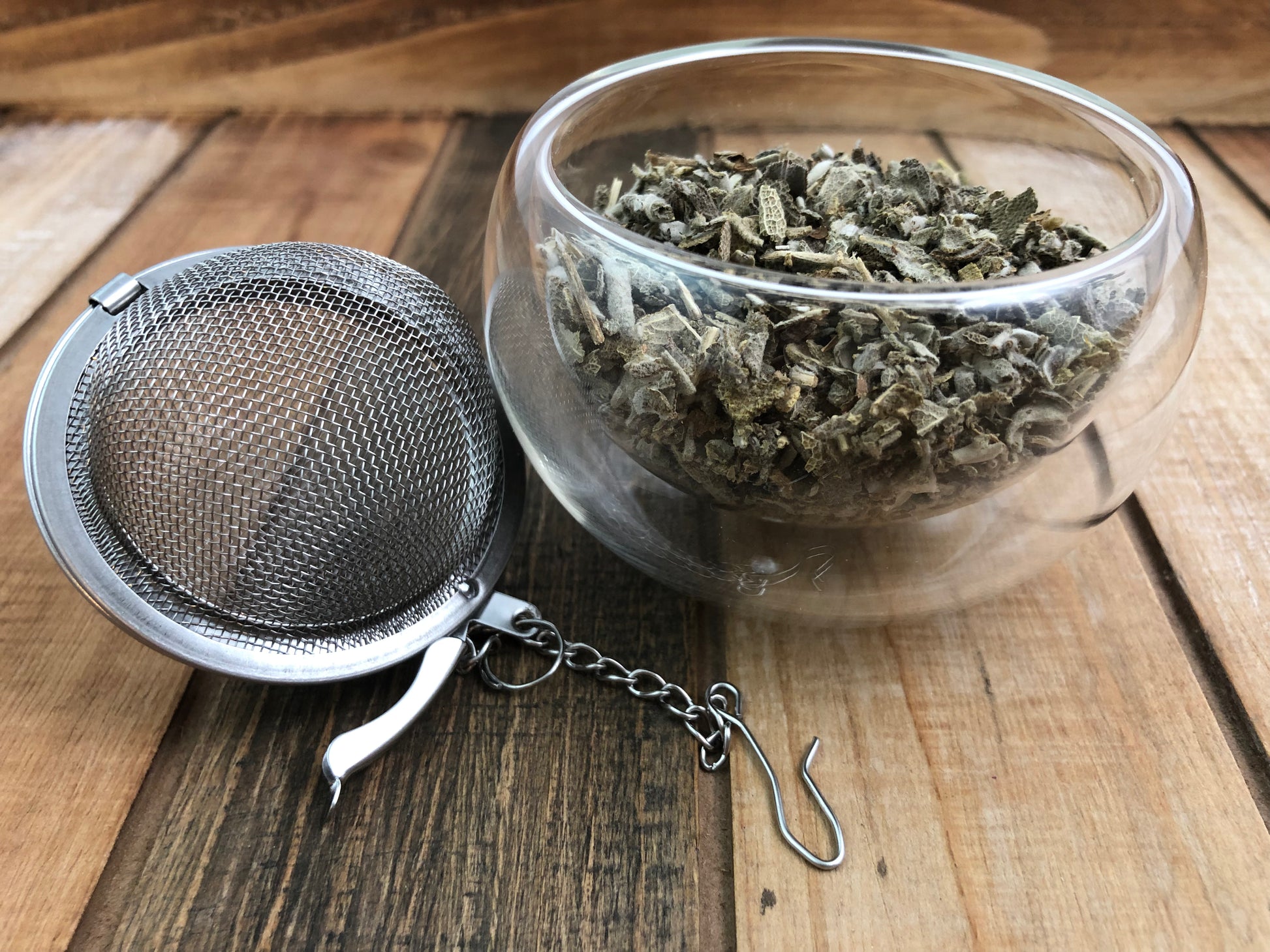 dried sage leaves in a clear glass bowl next to a tea ball infuser on wooden surface