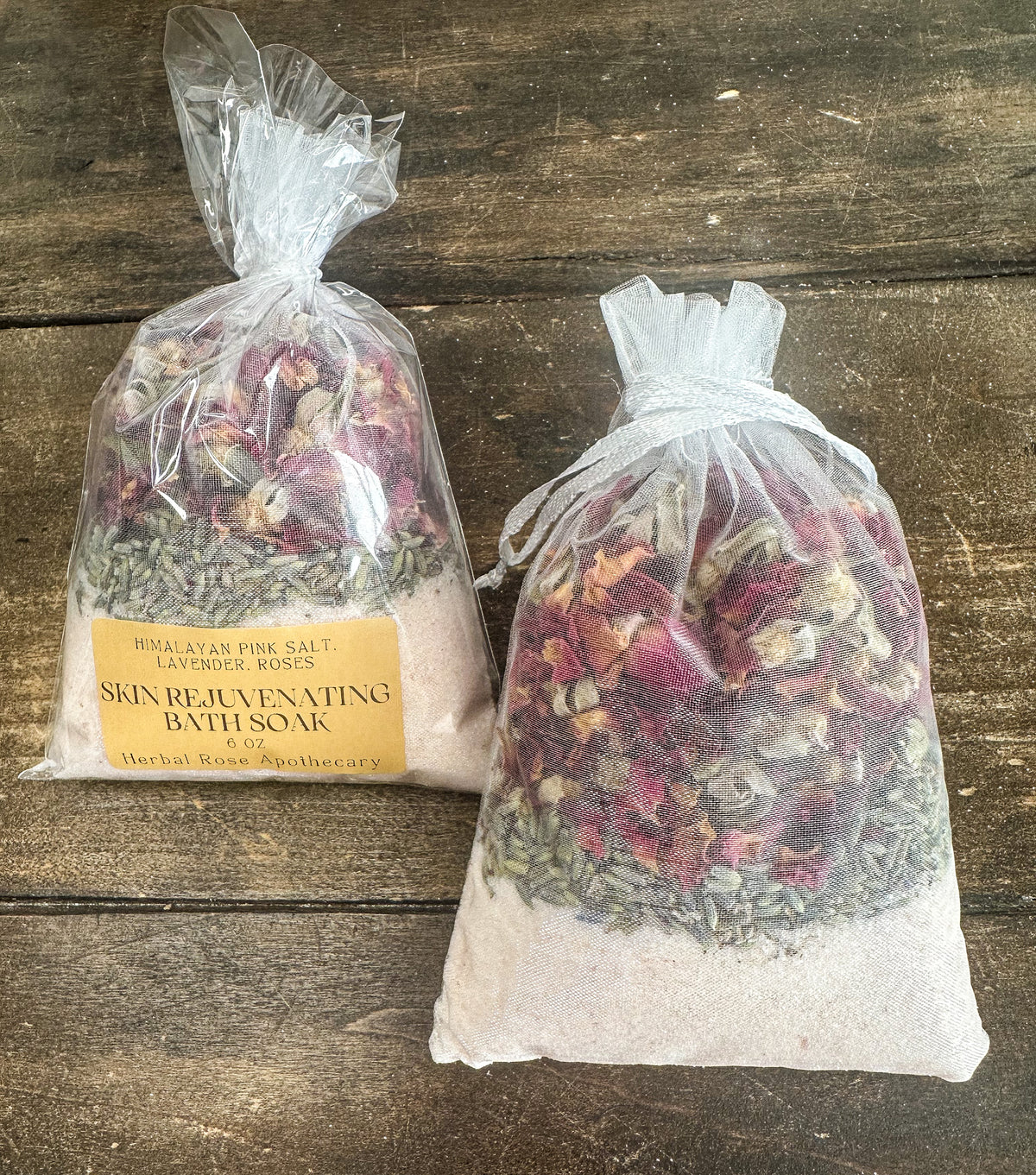 skin rejuvenating bath soak packaged in a clear bag and labeled; and one bath soak unpackaged lying on a wooden surface from herbal rose apothecary