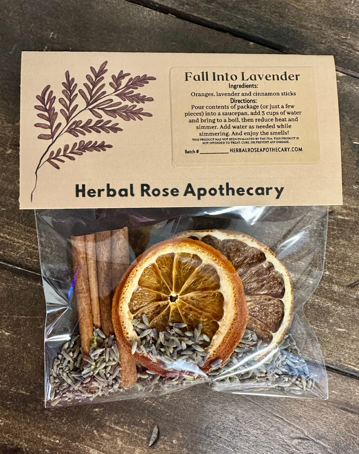 A clear plastic bag containing dried orange slices, cinnamon sticks, and lavender buds sits on a dark wooden surface beneath a kraft paper label. The label reads 'Fall Into Lavender' and 'Herbal Rose Apothecary'.