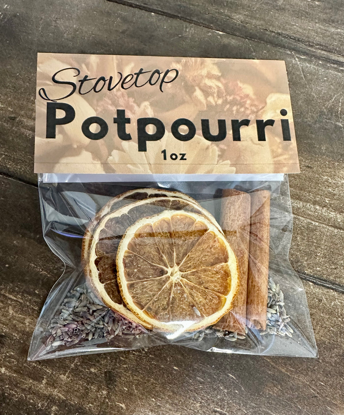 A 1 oz clear plastic bag of Stovetop Potpourri containing dried orange slices, lavender buds, and a cinnamon stick, with a brown paper label on a dark wood background.