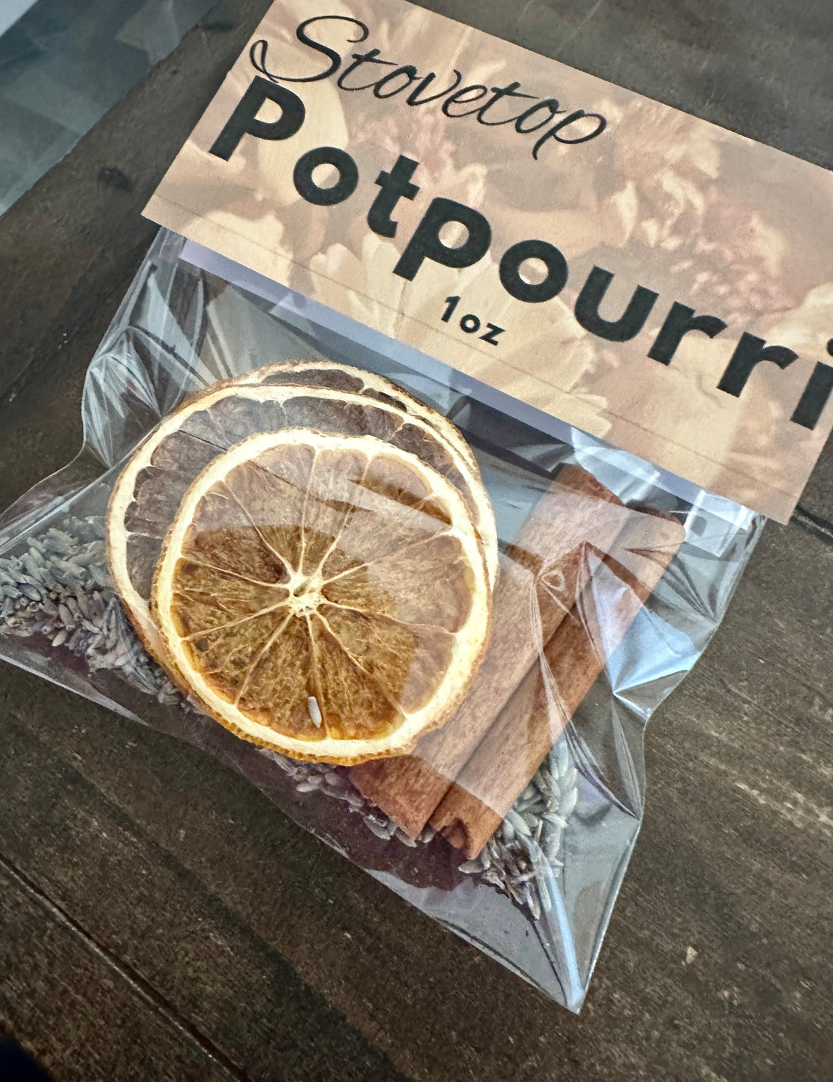 A 1 oz bag of Stovetop Potpourri containing dried orange slices, cinnamon sticks, and lavender buds, sealed in a clear plastic bag with a brown paper label, resting on a dark wooden surface.
