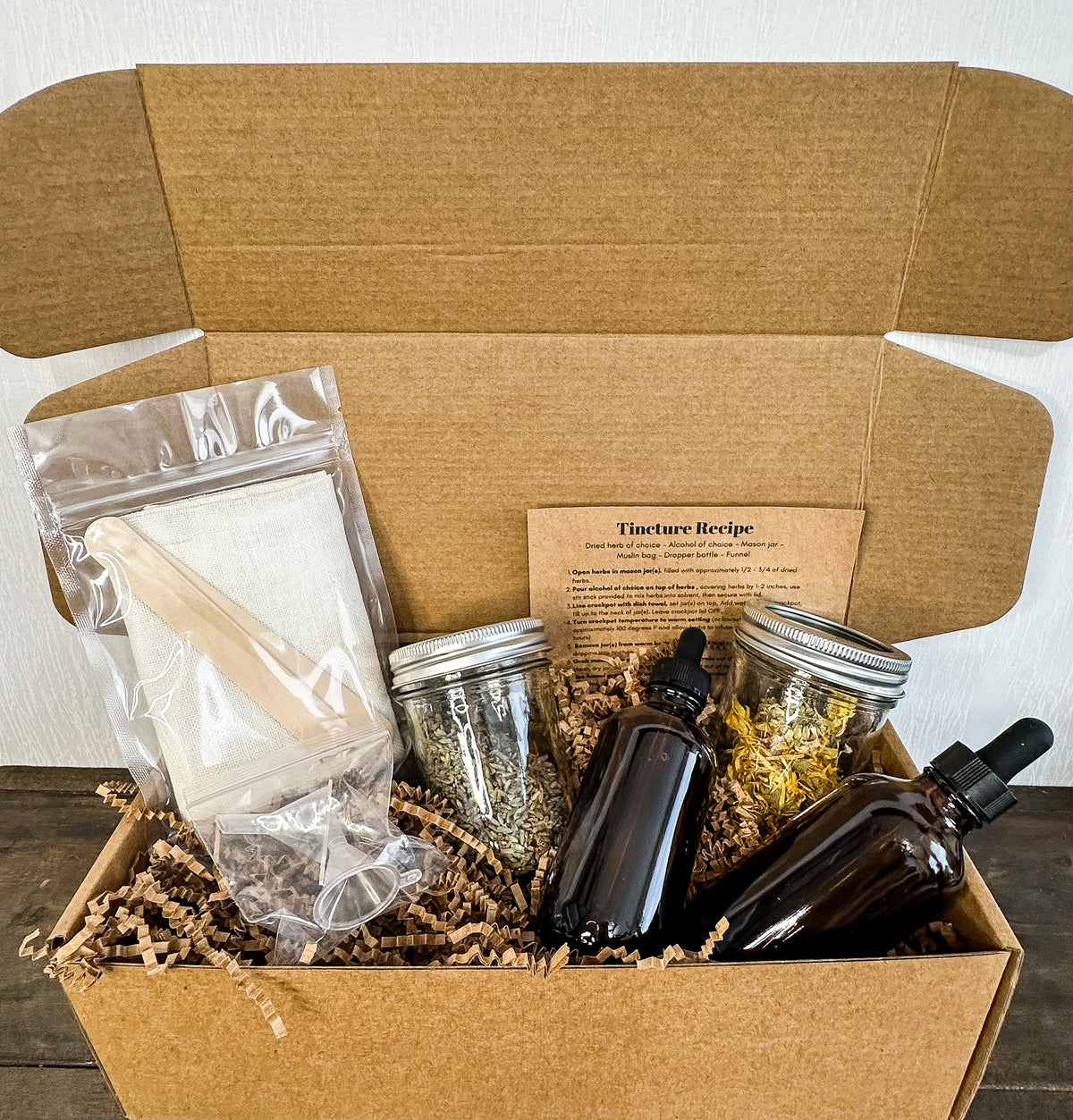 DIY tincture kit box opened to see included products; 2 herb jars, 2 amber dropper bottles, 2 funnels, a recipe card and  2 cheesecloths and stir sticks laid out on a wooden surface