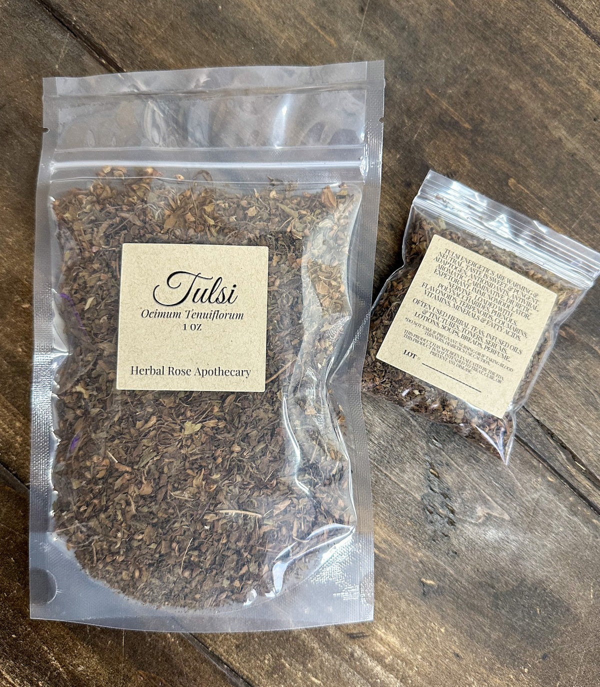 two packages of dried tulsi leaves in clear resealable bags from herbal rose apothecary on a wooden surface