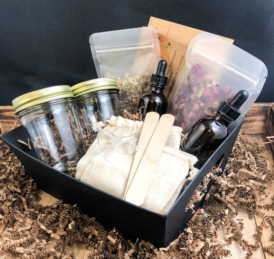 image of 2 herb tincture kit in a black basket with Kraft crinkle paper surrounding basket, sitting on wooden table with black background. basket includes 2 cards, 2 bags of dried herbs, 2 amber dropper bottles, 2 mason jars, 2 tongue depressors, 2 muslin bags