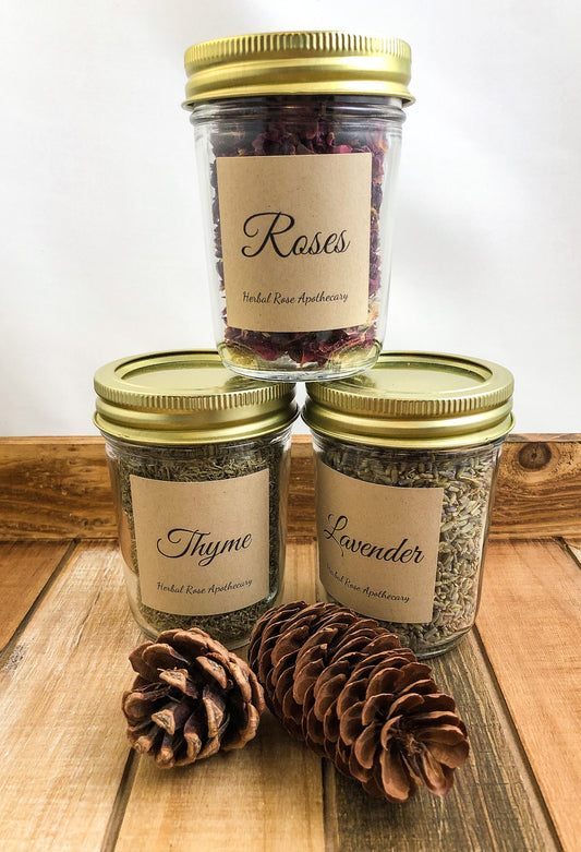 3 mason jars filled with dried herbs with Kraft colored labels with herb names stacked with one on top of two others, with 2 pinecones in forefront, white background and wooden table
