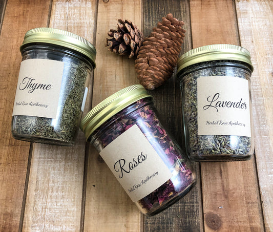 three glass mason jars filled with dried herbs with Kraft colored labels indicating herb with 2 pinecones in image with a wooden background