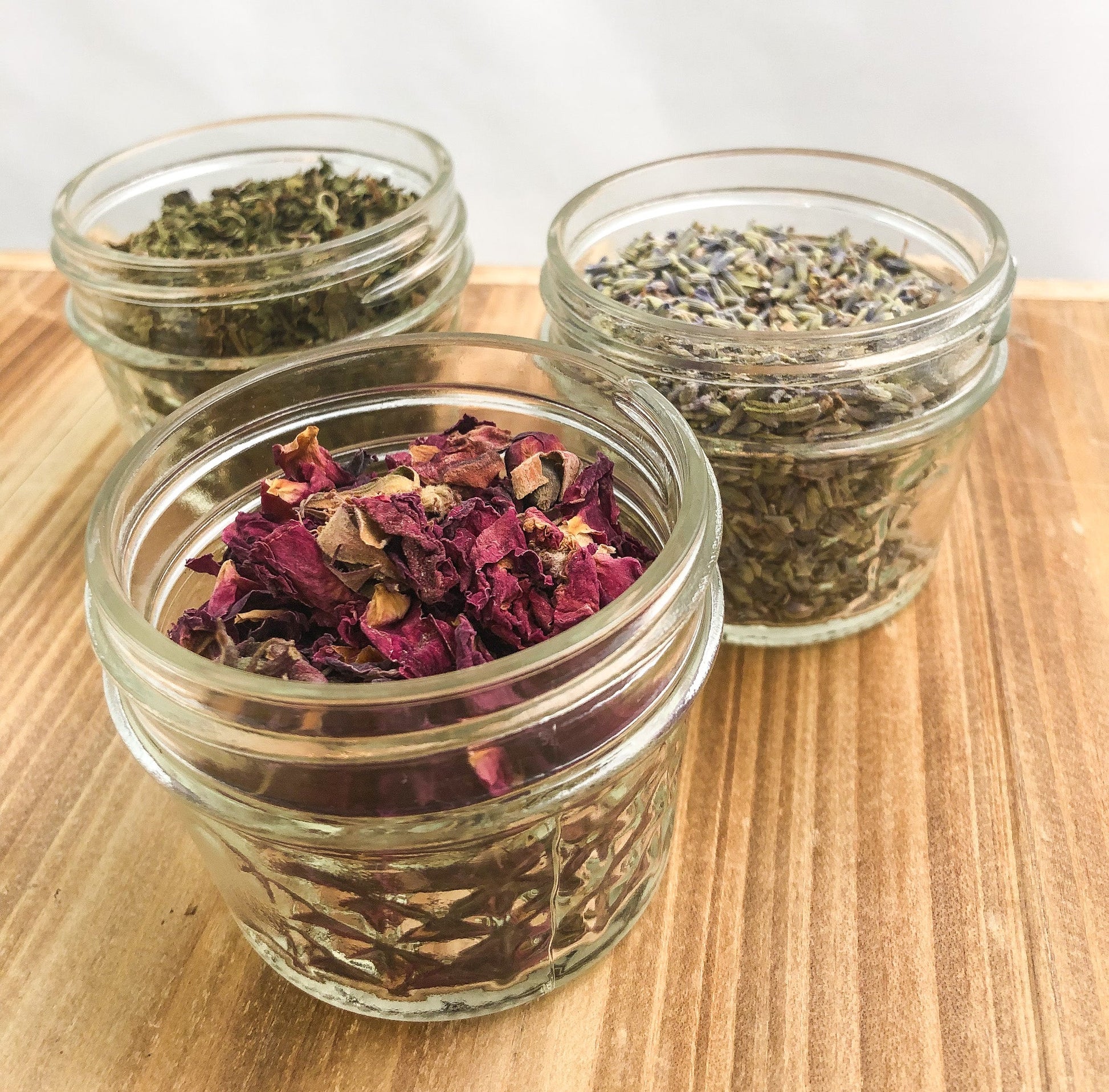 an angled image of 3 mini glass jars without lids filled with dried herbs on a wooden table top with a white background