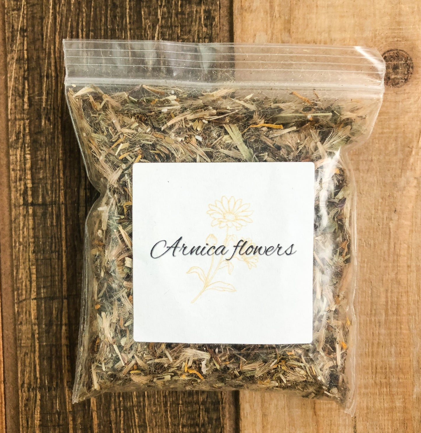 dried arnica flowers in a small clear plastic bag with a white label on front and wooden background
