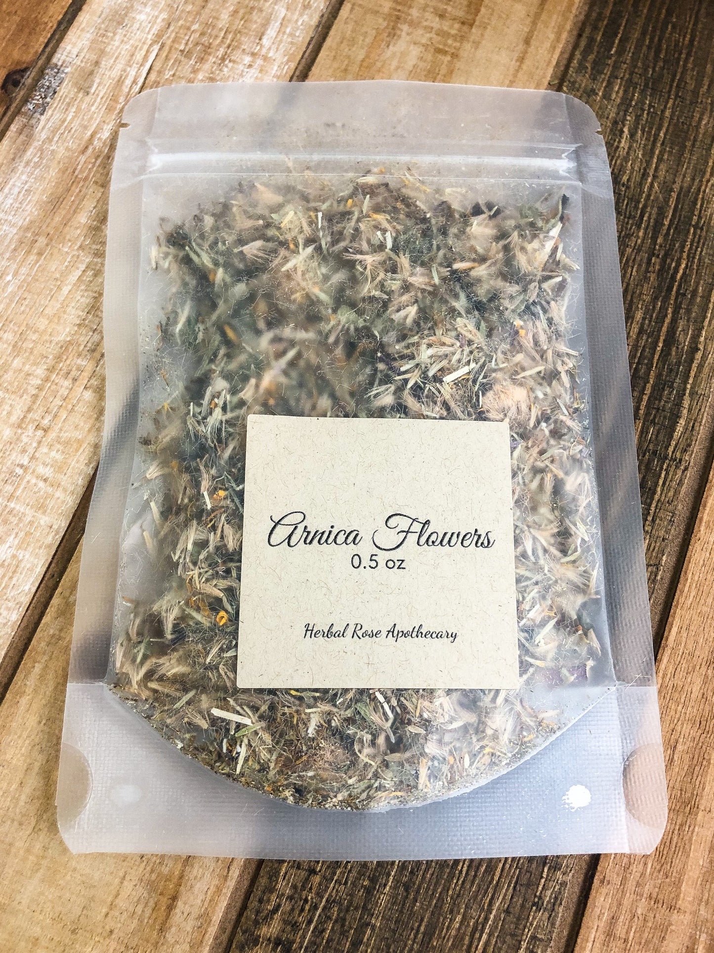 dried arnica flowers in clear plastic bag with Kraft colored label on bag indicating herb name and size of 0.5oz with a wooden background