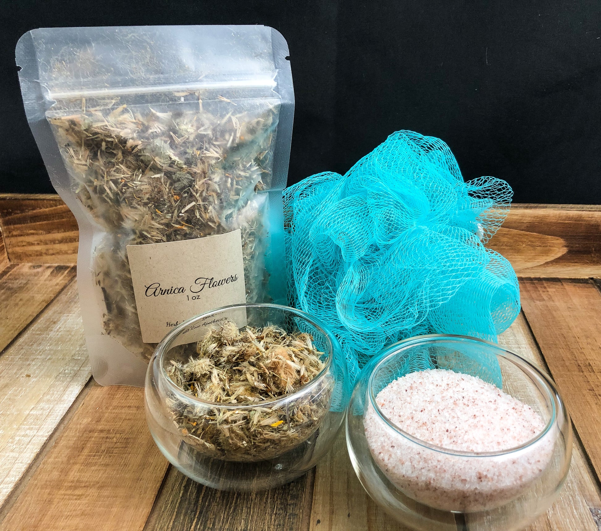 dried arnica flowers in a 1oz bag next to a bath sponge and two clear bowls of arnica flowers and pink Himalayan salt all on a wooden tray with black background