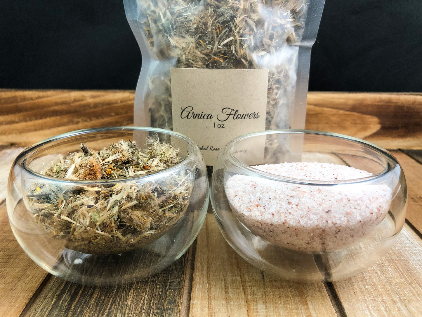 upclose image of two clear bowls of arnica flowers and Himalayan salt with dried arnica flowers in a 1oz bag behind them, all on a wooden tray with black background