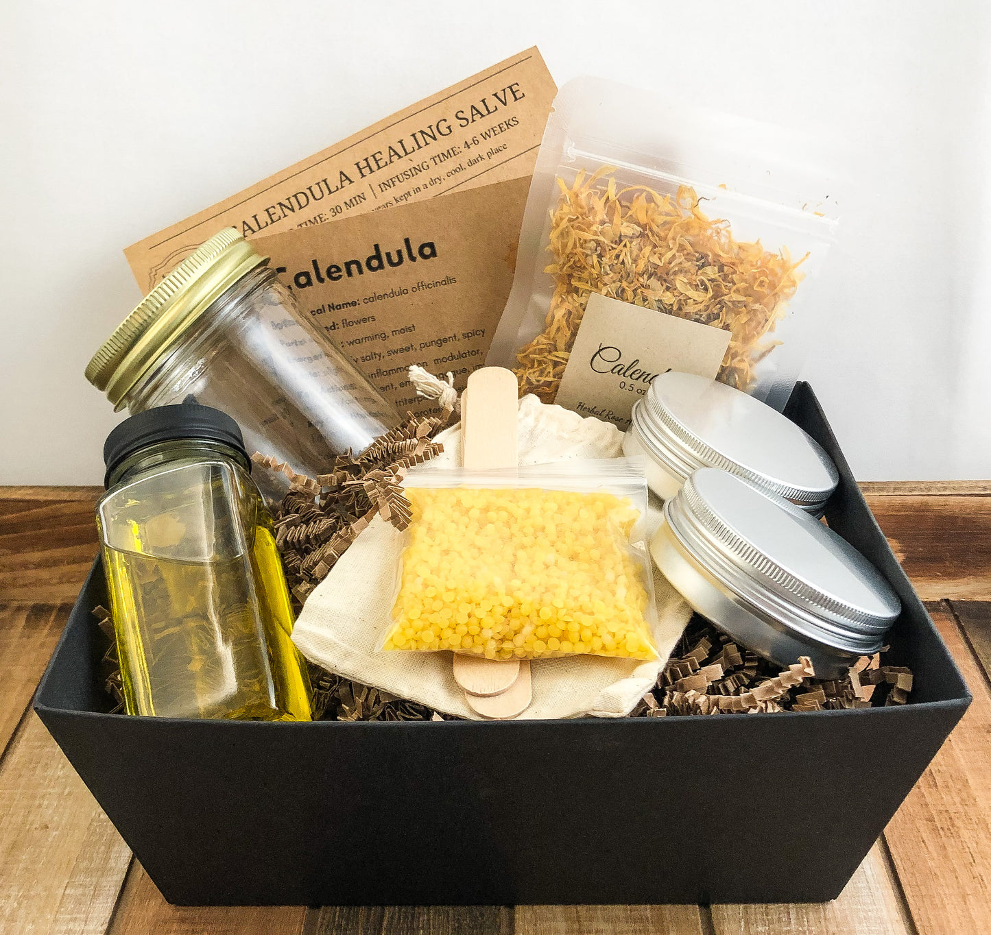 calendula salve kit in a black basket with with white background. Kit includes directions, monograph, 2 metal tins, dried calendula, mason jar, beeswax, wooden sticks, and muslin bag