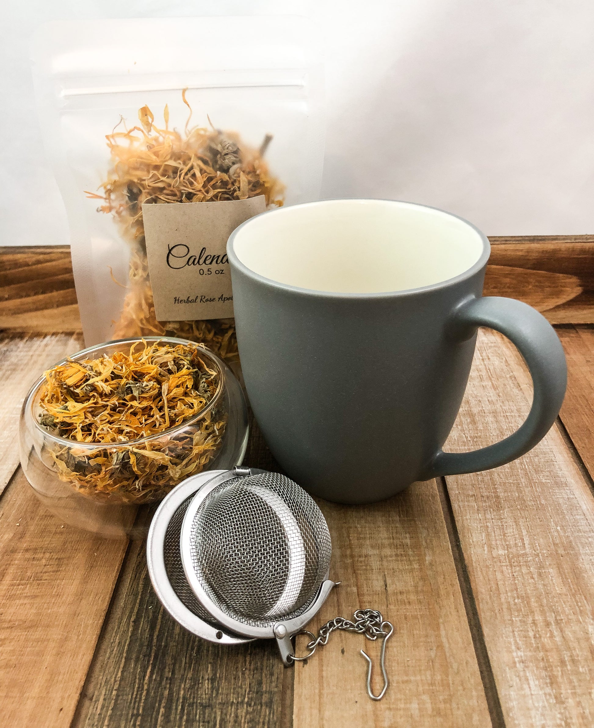 image with a clear small bowl of dried calendula flowers and mesh strainer in the forefront, a mug to the right of the image grey on the outside and white on the inside, and a bag of 0.5oz dried calendula in a clear plastic bag behind the mug, with a whitish background and items sitting on a wooden table