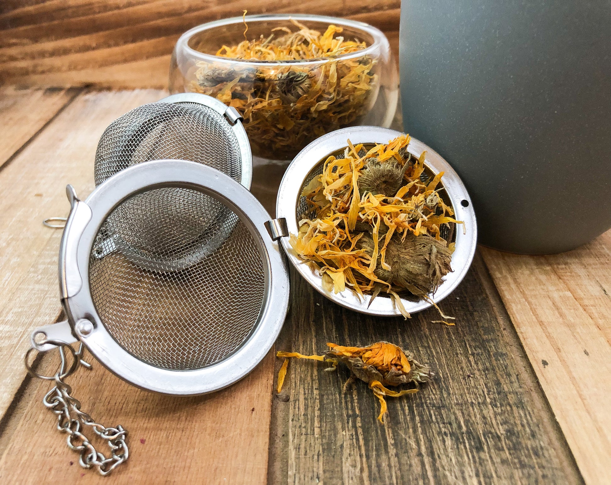 dried calendula flowers in an open mesh tea strainer with some flowers outside of strainer, another mesh strainer behind open one, a clear small glass bowl with dried calendula flowers sitting behind and next to a grey mug with a wooden background