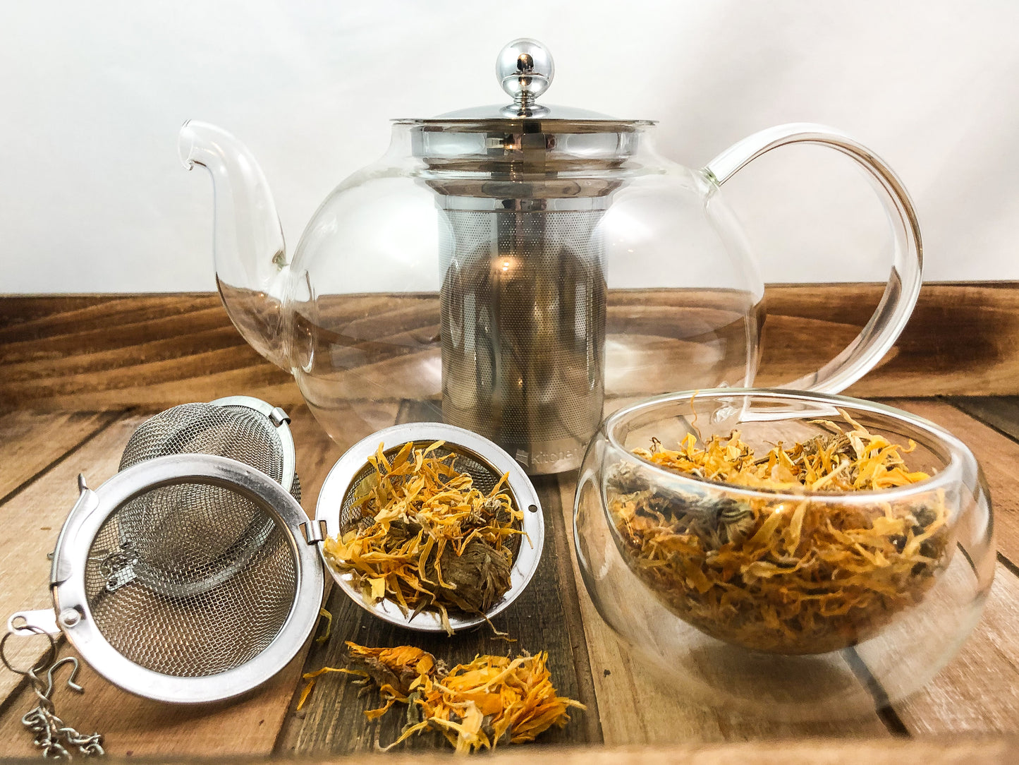 clear teapot with a clear small glass bowl full of dried calendula flowers in forefront, next to two mesh tea strainers, one being open with dried calendula flowers inside and falling out, with a white background and wooden table