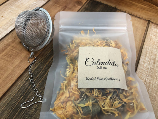dried calendula flowers in a clear plastic bag with a Kraft colored label on front of bag indicating 0.5oz in size and herb name, next to a mesh tea strainer on a wooden background