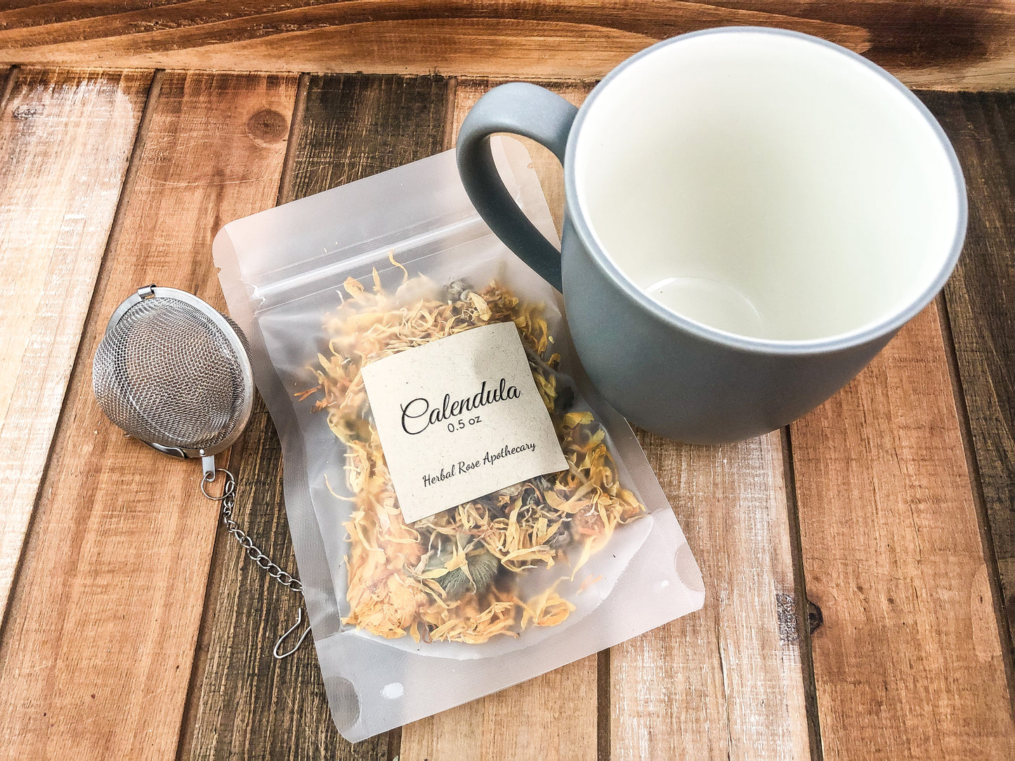 image is looking down into mug on a wooden background with a mug that is grey on outside and white inside, mesh tea strainer and a bag of dried calendula flowers in a 0.5oz size bag 