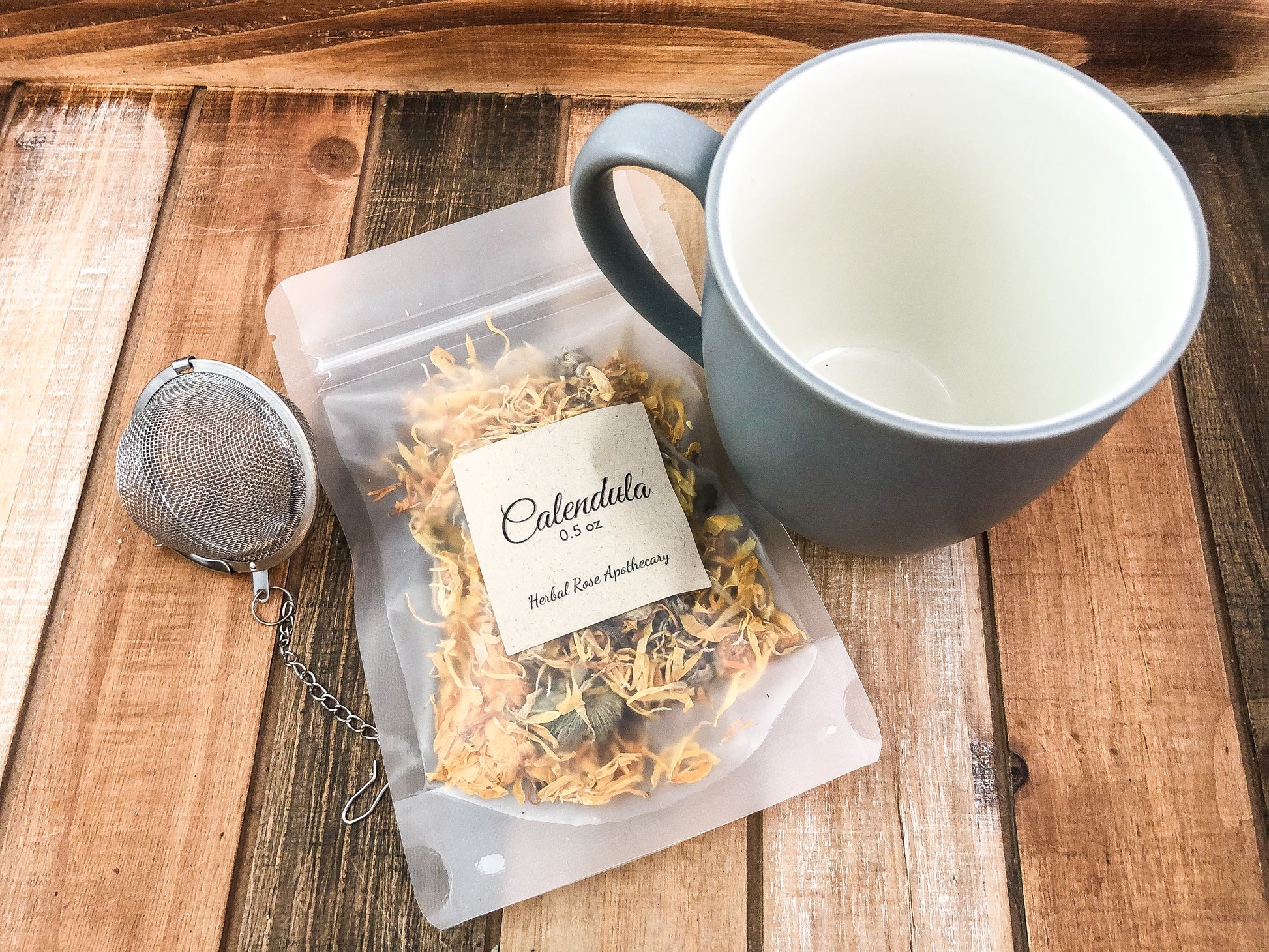 image is looking down into mug on a wooden background with a mug that is grey on outside and white inside, mesh tea strainer and a bag of dried calendula flowers in a 0.5oz size bag 