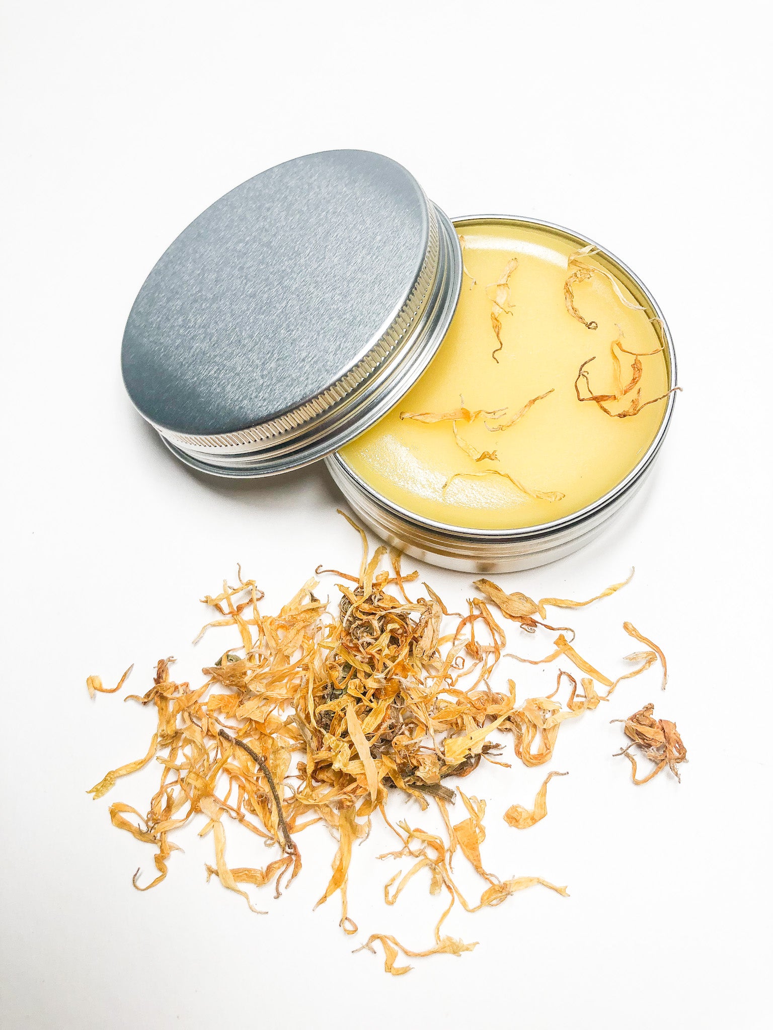 calendula salve in metal tin with dried calendula flowers scattered on top of salve and in forefront all with a white background