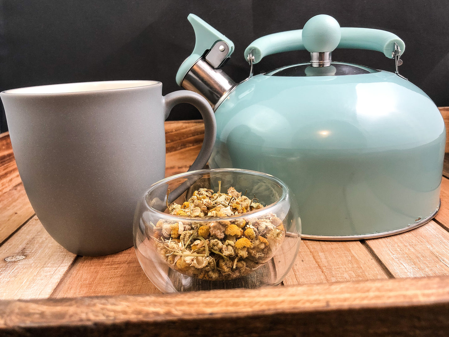 clear small bowl of dried chamomile in forefront with grey mug to left and teal colored tea pot to the right on a wooden table with a black background